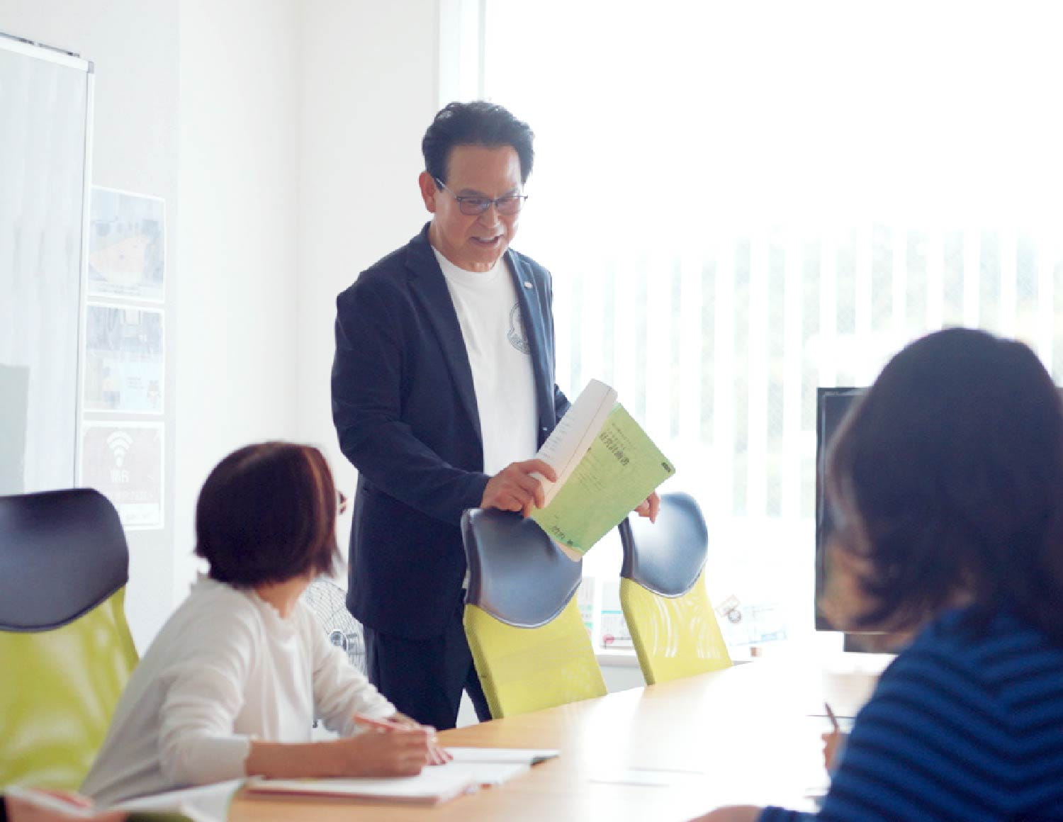 スーツの男性が立って話し、会議テーブルを囲む複数の社員が聴いている様子