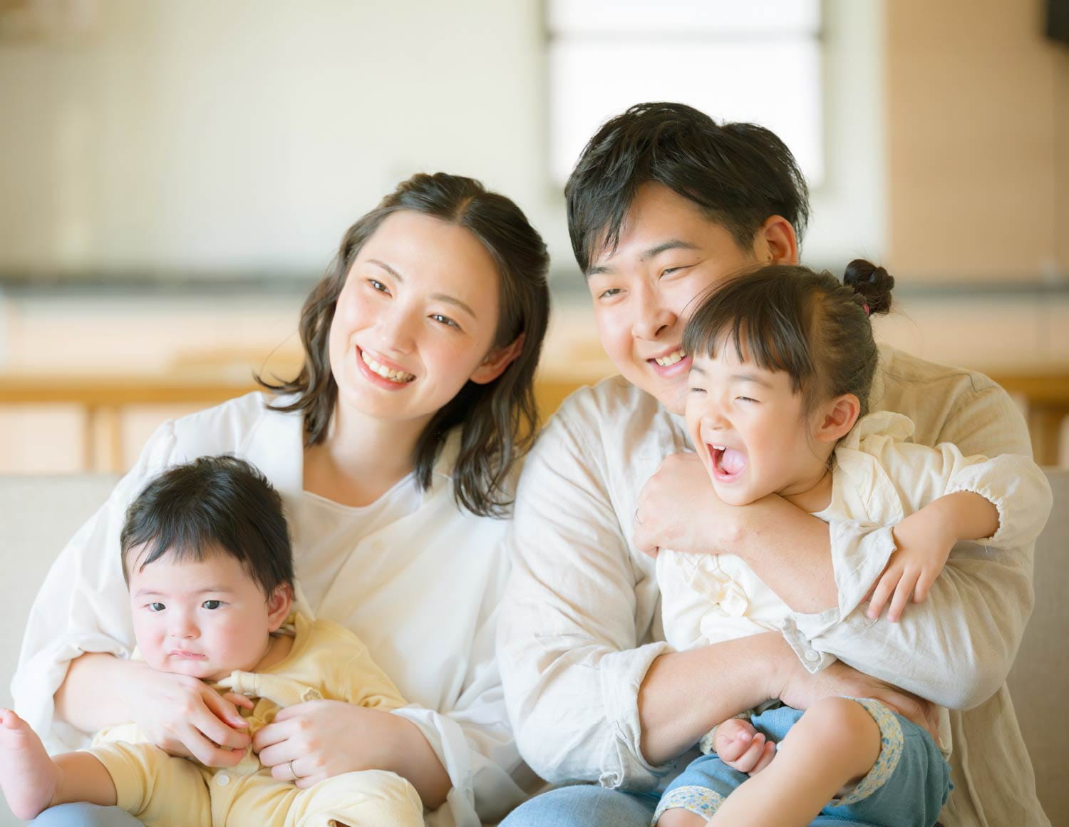 明るい室内で、父母と幼い二人の子どもが笑い合い寄り添う家族の様子