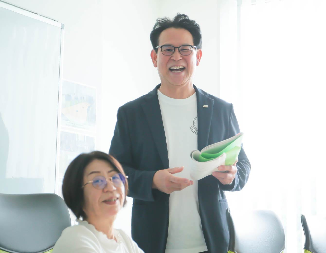 オフィスで微笑む男女二人が、明るい室内で肩を並べてこちらを向いている