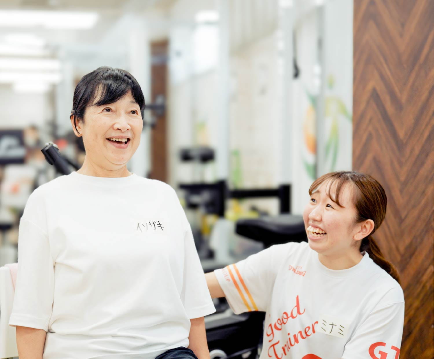白いロゴ入りシャツの女性トレーナーが、お客様と向き合い笑顔で話している様子