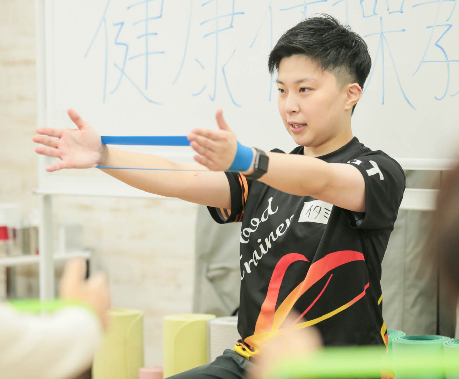 黒とオレンジのウェアの男性が、青いチューブを引き伸ばし健康体操の前で実演している