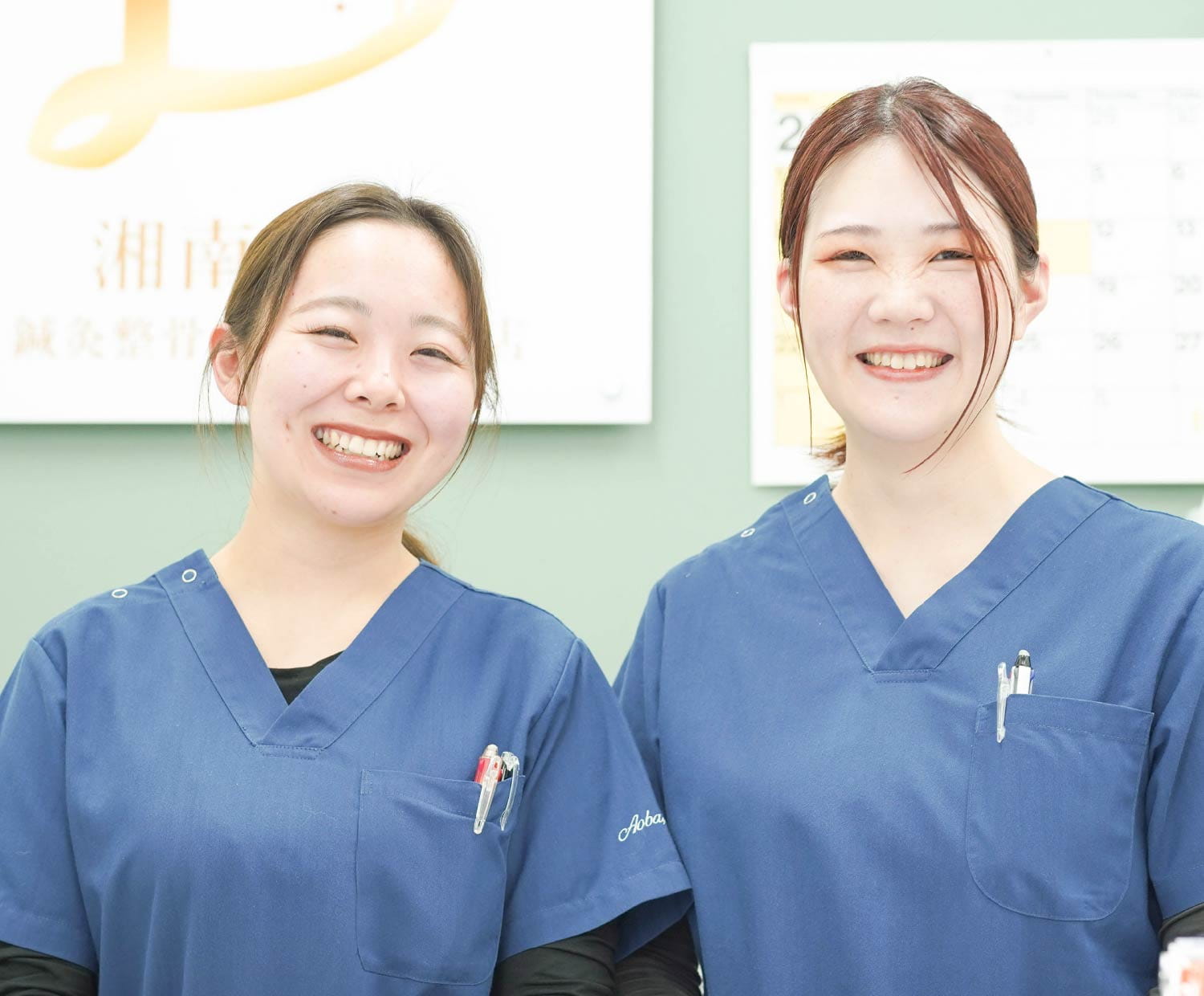 青色スクラブを着た女性スタッフ二人が、明るい院内で並んで笑顔を見せている