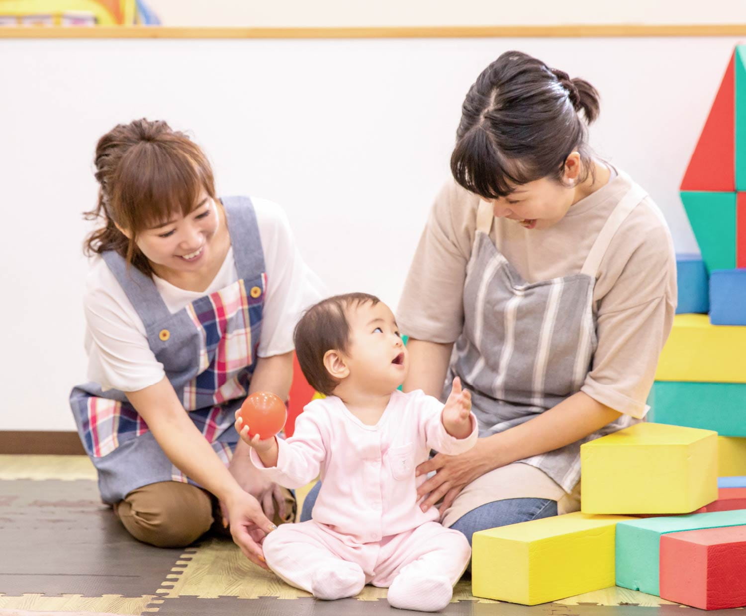 カラフルなマットの上で赤ちゃんを囲み、女性スタッフ二人が笑顔で見守っている様子
