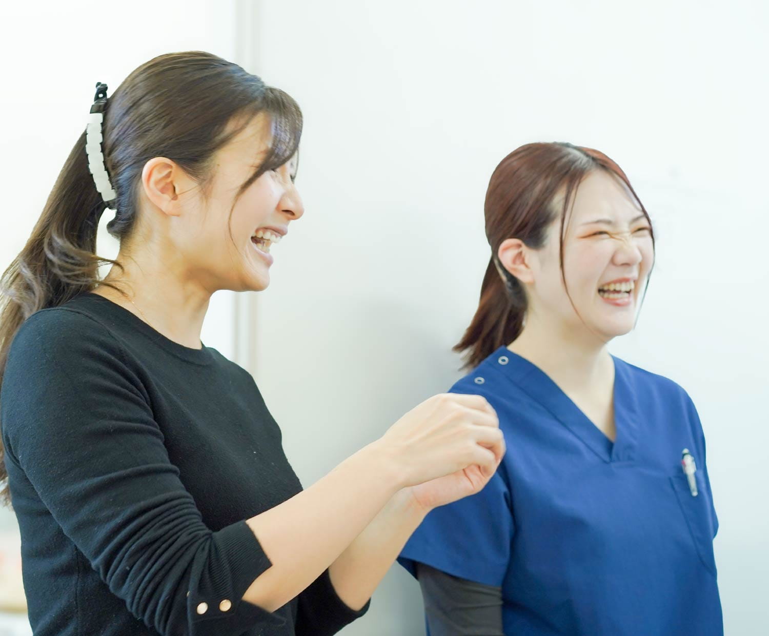 黒いトップスの女性とスクラブ姿の同僚が、明るい室内で笑い合いながら向き合っている