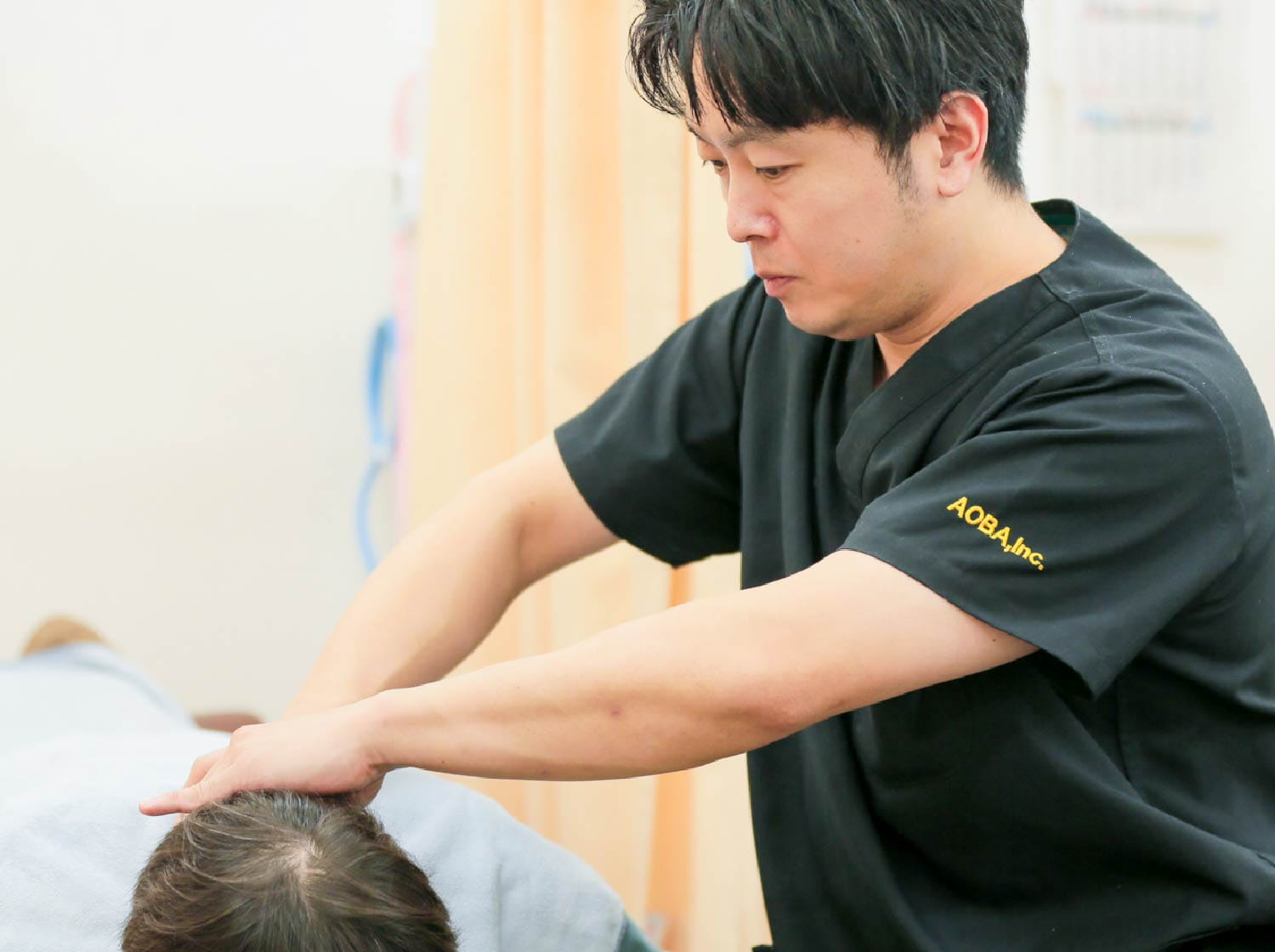 黒いスクラブの治療者が、うつ伏せの患者の頭部に施術の手を置いている様子